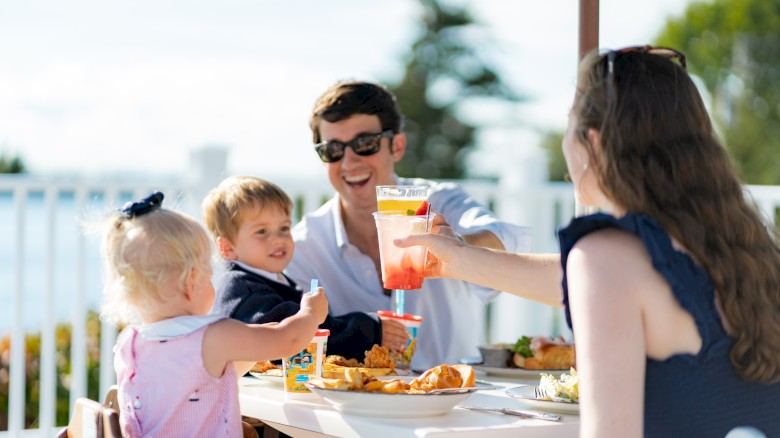 A family of four enjoys a meal outdoors, smiling and raising drinks, with food on the table and a scenic background.