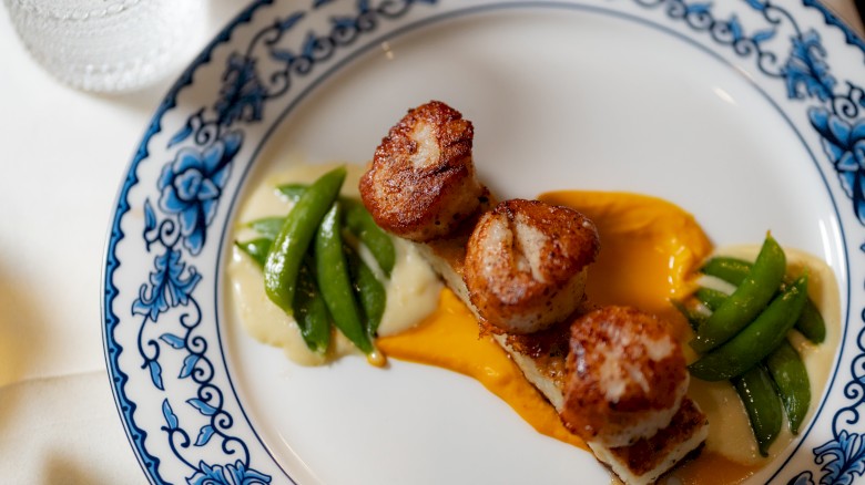 Seared scallops placed on a decorative plate with sauces and green vegetables, probably snap peas, garnished beautifully.