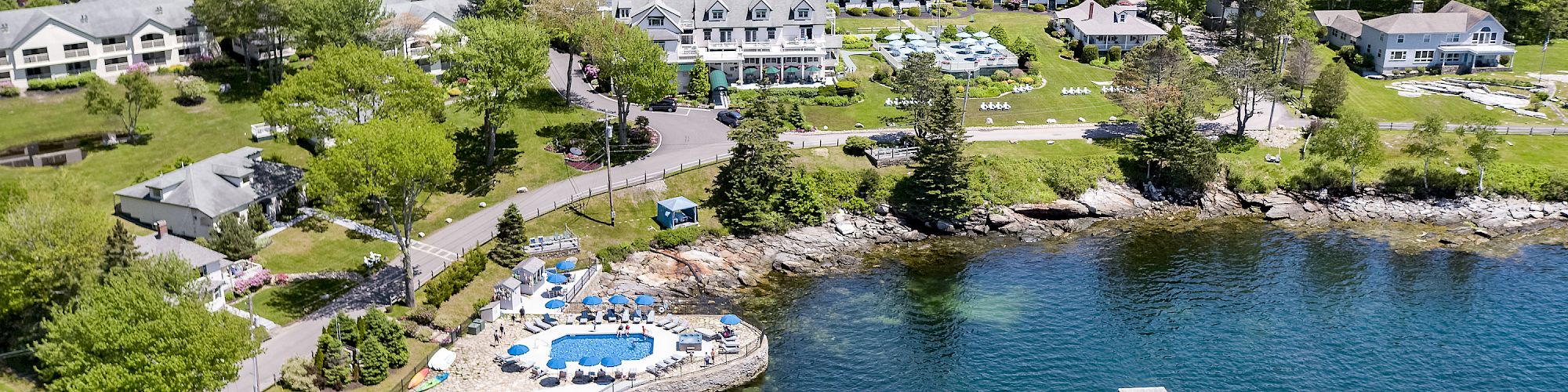 Aerial view of a waterfront resort with multiple buildings, a dock, boats, and a swimming pool, surrounded by lush greenery and water.