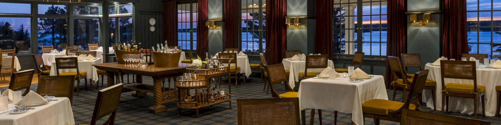 A dimly lit, elegant restaurant with white tablecloths, folded napkins, and fine tableware, featuring large windows and hanging light fixtures.