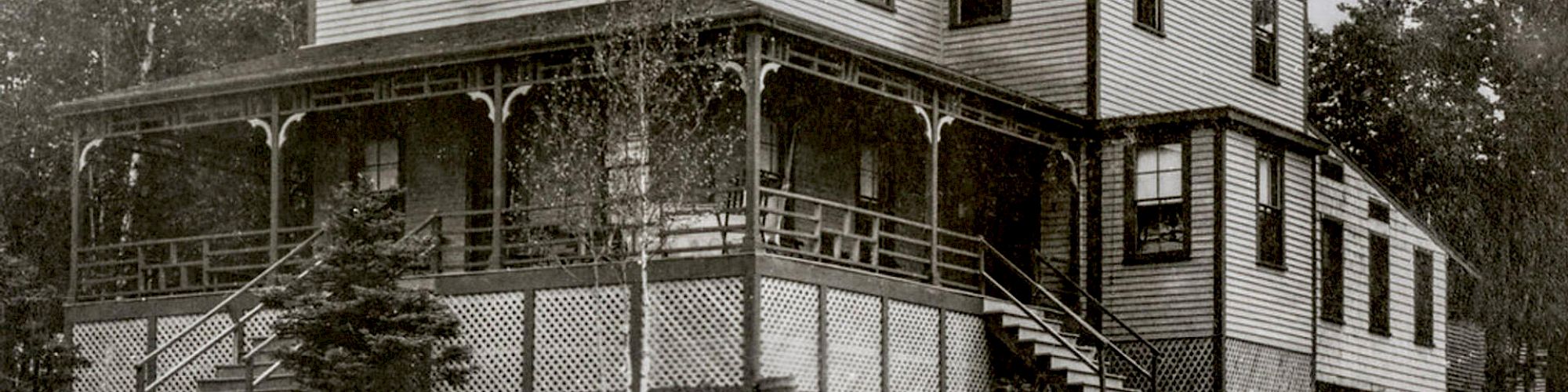 An old, black-and-white photo of a large, two-story house with a wraparound porch, multiple windows, and surrounding trees.