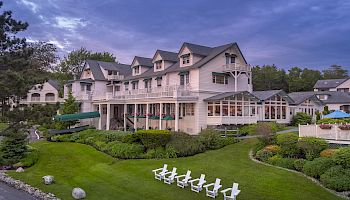 A large, elegant building with multiple dormer windows, surrounded by lush greenery and white chairs on the lawn under a picturesque sky.