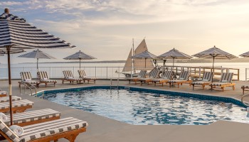 The image shows a serene poolside area with striped lounge chairs and umbrellas overlooking calm waters and a sailboat, under a partly cloudy sky.