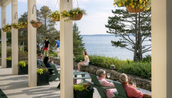 People sitting on Adirondack chairs on a porch overlooking a scenic waterfront, with hanging flower baskets and trees in the background, enjoying the view.