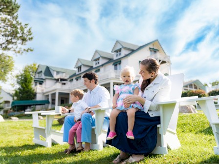 A family sitting on outdoor chairs in front of a large house, enjoying a sunny day on the lawn, with two parents and two children.