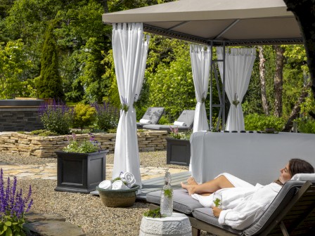 A person is relaxing on a lounge chair under a canopy in a garden, surrounded by greenery, flowers, and other lounge seating areas.