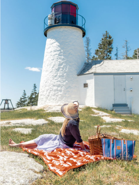 A person sits on a picnic blanket with a wicker basket near a white lighthouse under a clear blue sky and trees in the background.