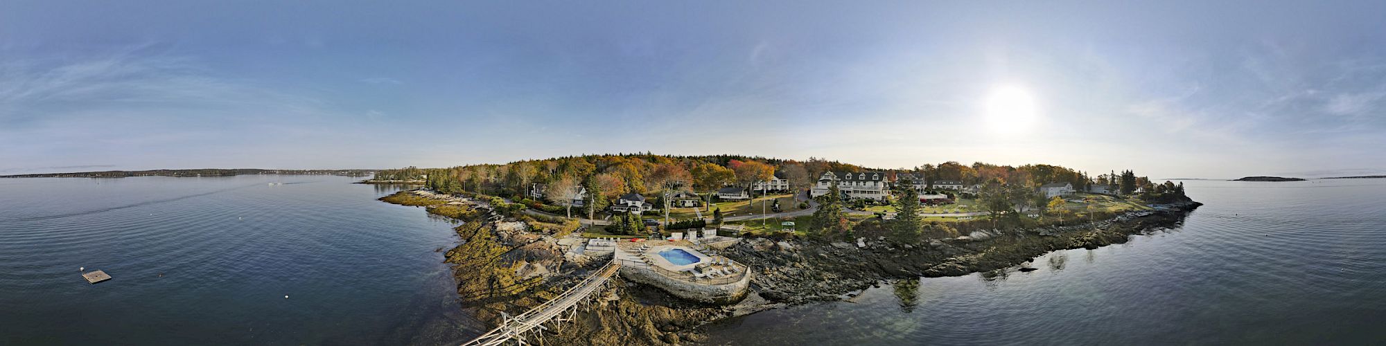 A panoramic view of a small island with a pier, various buildings, and a pool, surrounded by calm water and under a clear sky at sunset.