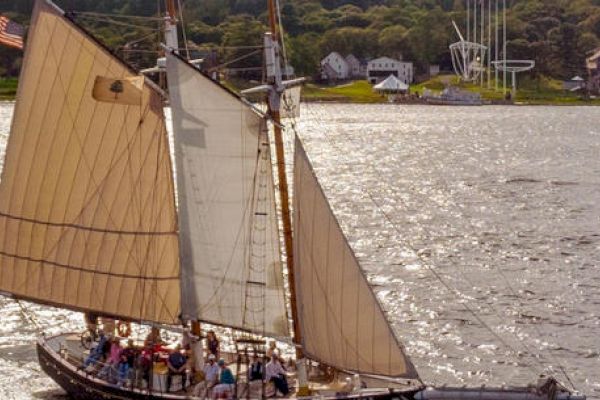 A sailboat with tan sails and multiple people onboard sailing on a body of water near a shoreline with buildings and trees in the background.