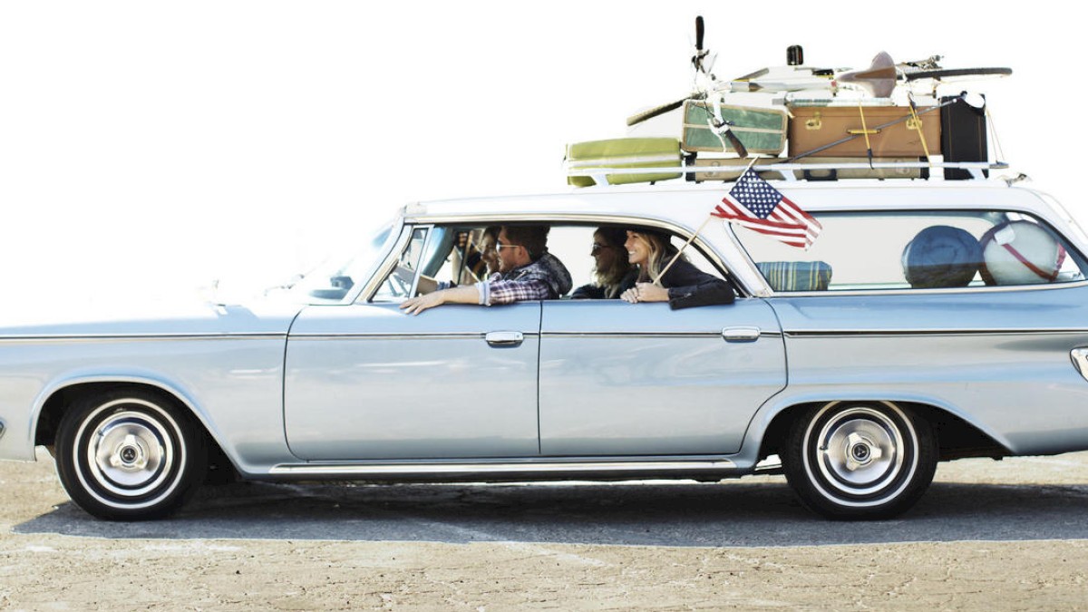 A vintage car with roof luggage and an American flag, carrying four people, parked on a sandy area.