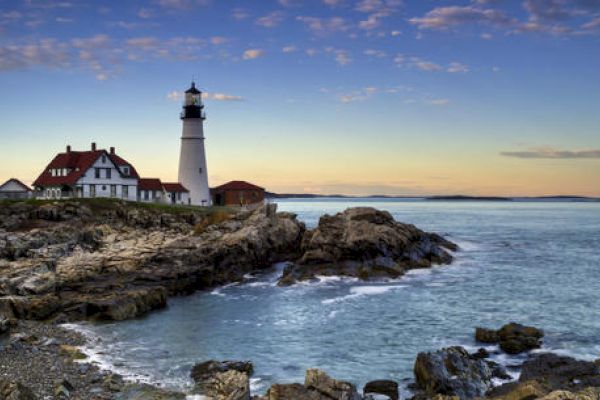 A lighthouse stands on a rocky coastline, with surrounding buildings and an expansive ocean under a calm, partly cloudy sky at sunset.