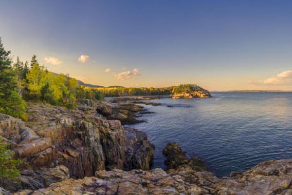 Rocky coastal landscape with cliffs, trees, and calm sea under a clear blue sky. Sunlight illuminates the scene, highlighting natural beauty.