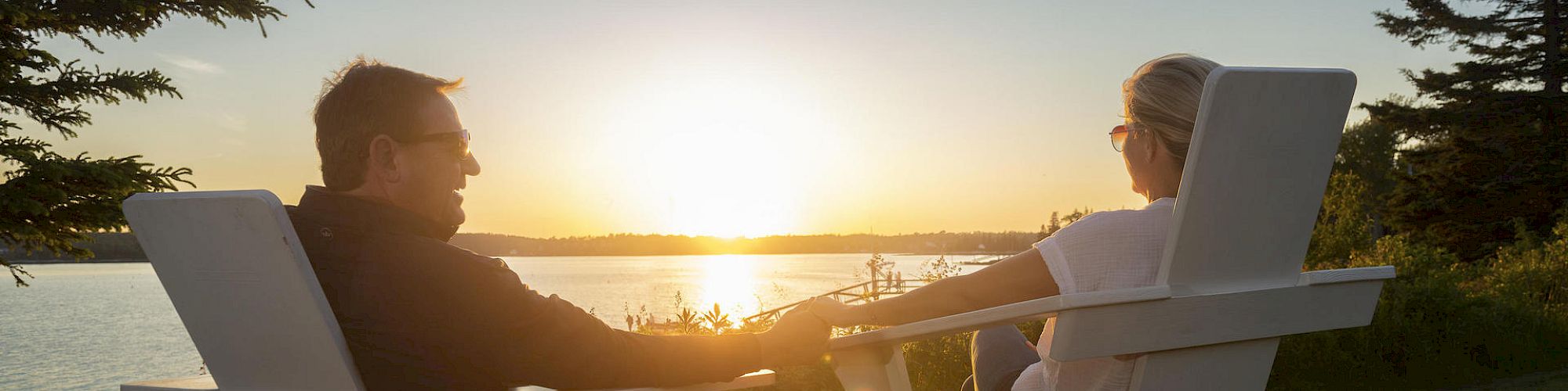 Two people are sitting in Adirondack chairs, holding hands, and watching the sunset over a lake, surrounded by trees and greenery.