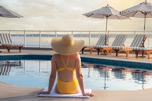 A person in a yellow swimsuit and wide-brim hat sits by a pool, facing away, with empty lounge chairs and umbrellas in the background at sunset.