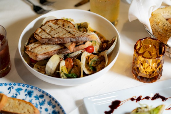 A bowl of seafood pasta with clams, tomatoes, and grilled bread, accompanied by a beverage and side dishes, is seen on a dining table.