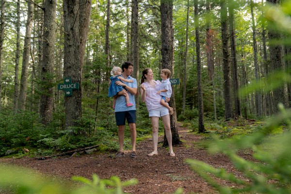 A family of four, standing in a forest, holding hands and smiling. The path splits, with signs saying 