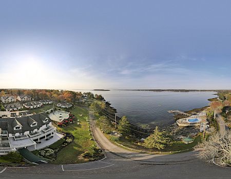 A panoramic view of a coastal resort featuring buildings, a swimming pool, trees with fall foliage, a body of water, and roads.