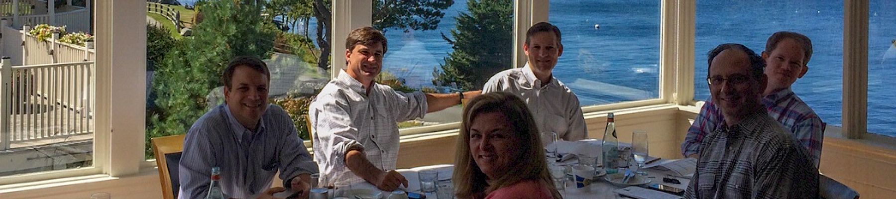 Six people sitting at a table with documents and drinks, overlooking a scenic ocean view from a large window.