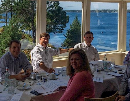 Six people sitting at a table with documents and drinks, overlooking a scenic ocean view from a large window.