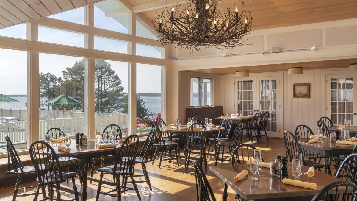 A cozy restaurant with wooden tables and chairs, large windows offering a scenic view of water and trees, and a rustic chandelier on the ceiling.