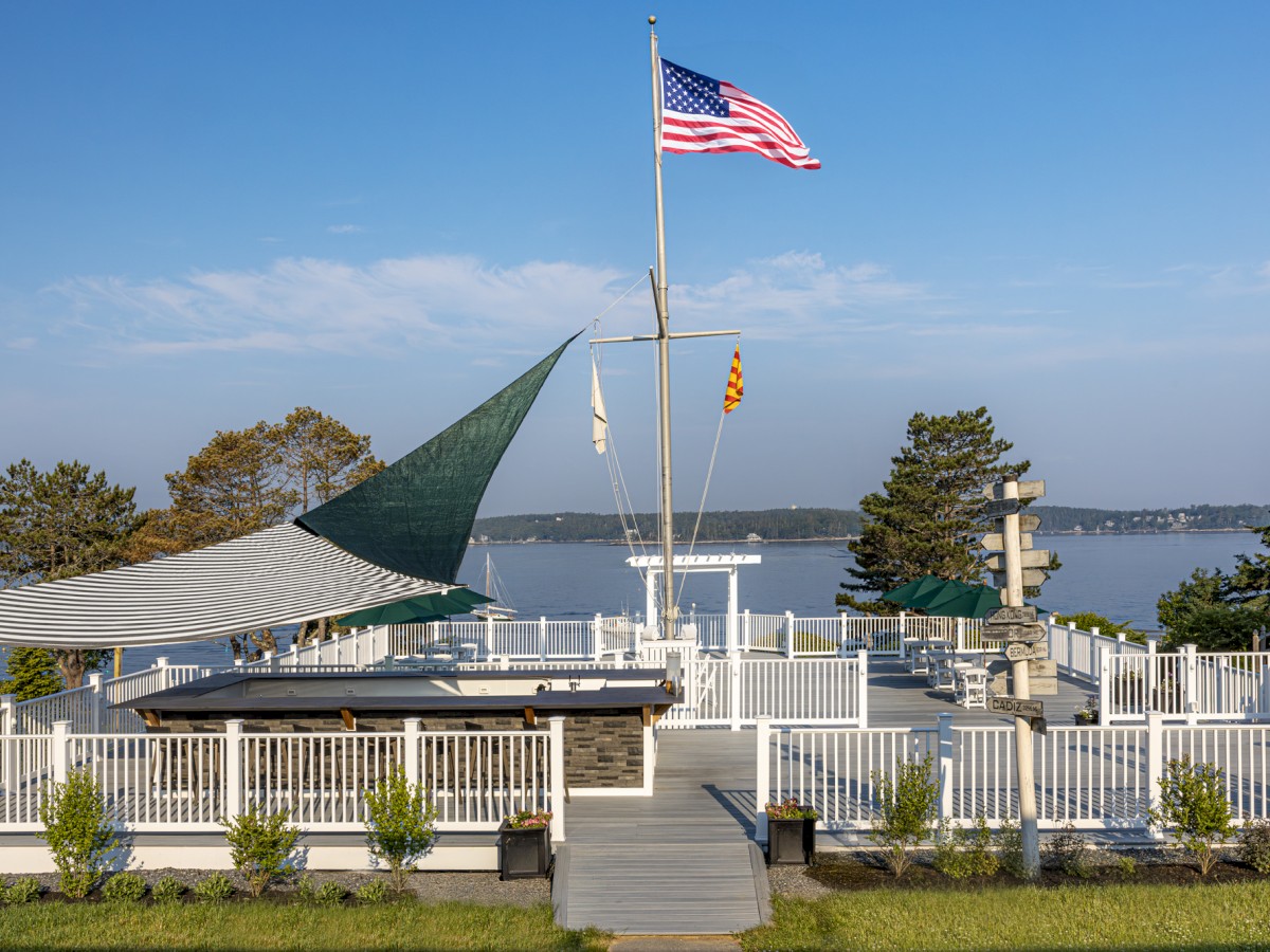 A deck with a flagpole, an American flag, and a scenic view of water and trees in the background, surrounded by a white fence, is pictured.