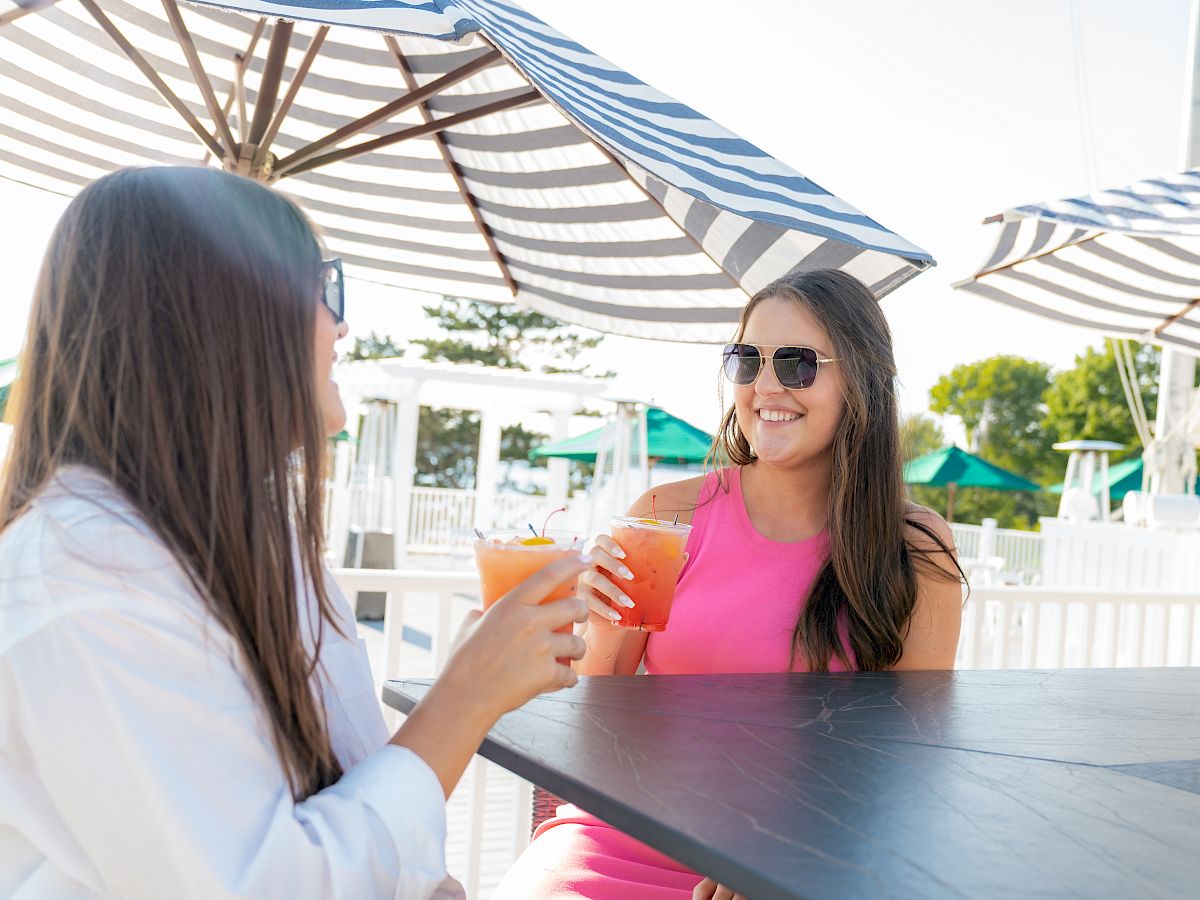 Two women are sitting outdoors under umbrellas, smiling and holding drinks, enjoying a sunny day together.