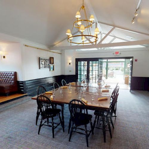 A spacious dining area with a large wooden table, black chairs, chandeliers, rock wall with a ship wheel, and leather booth seating.