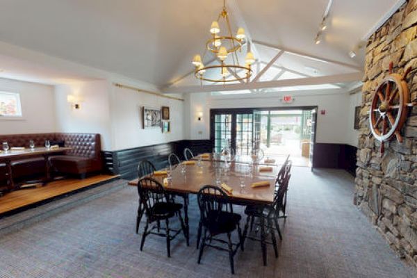 A dining room with a long table, black chairs, chandeliers, a stone wall with a ship wheel, and a booth seating area with framed artwork.