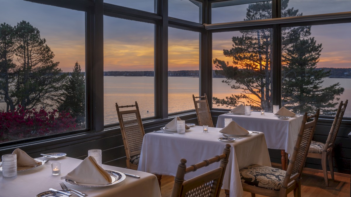 A cozy restaurant with tables set for dining, large windows offering a stunning sunset view over a body of water, and a few trees visible outside.