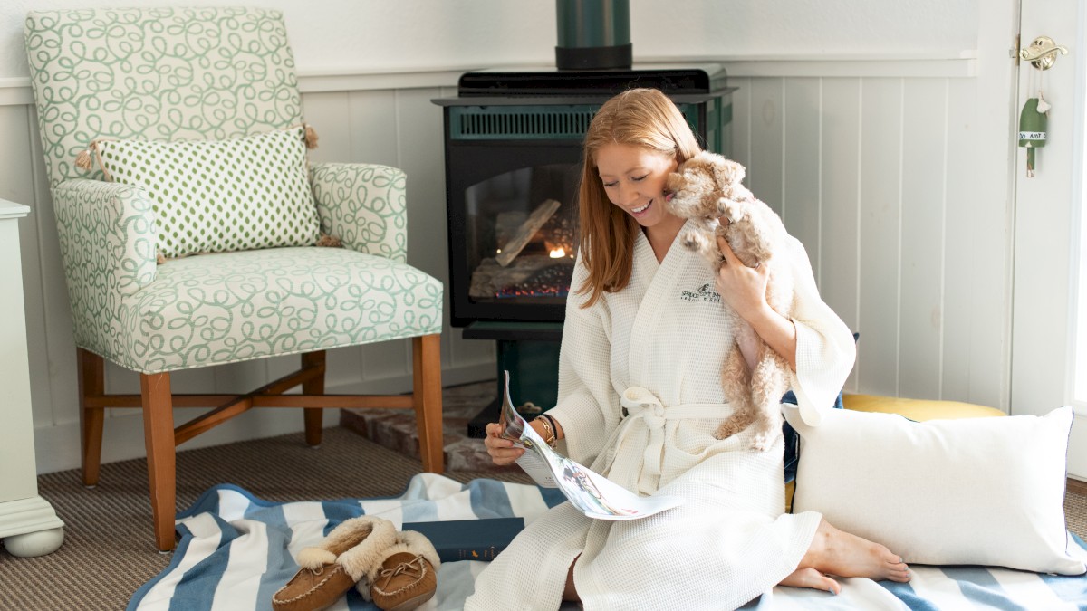 A woman in a white robe is sitting on a striped blanket, reading a magazine, and holding a dog with slippers nearby in a cozy room.