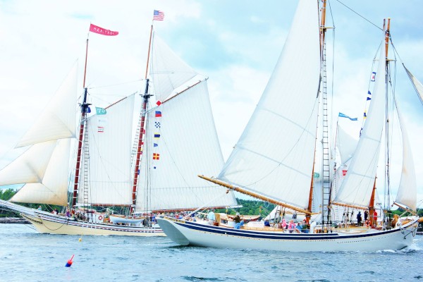 Two sailboats with white sails and flags are navigating on the water under a cloudy sky, with people aboard.