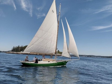 A sailboat with white sails is on a calm body of water with people on board, under a blue sky with wispy clouds in the distance by a small island.