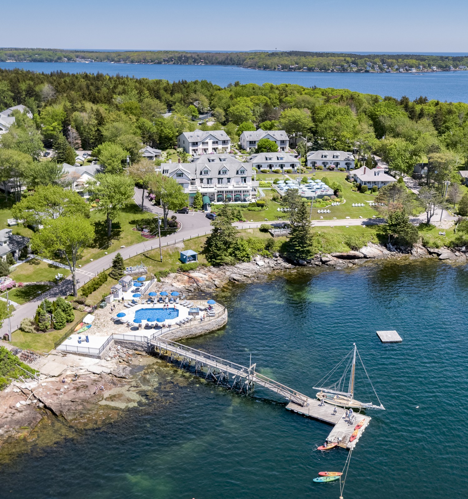 An aerial view of a lakeside resort with buildings, a pool area, green lawns, and a dock extending into the water with a small boat.