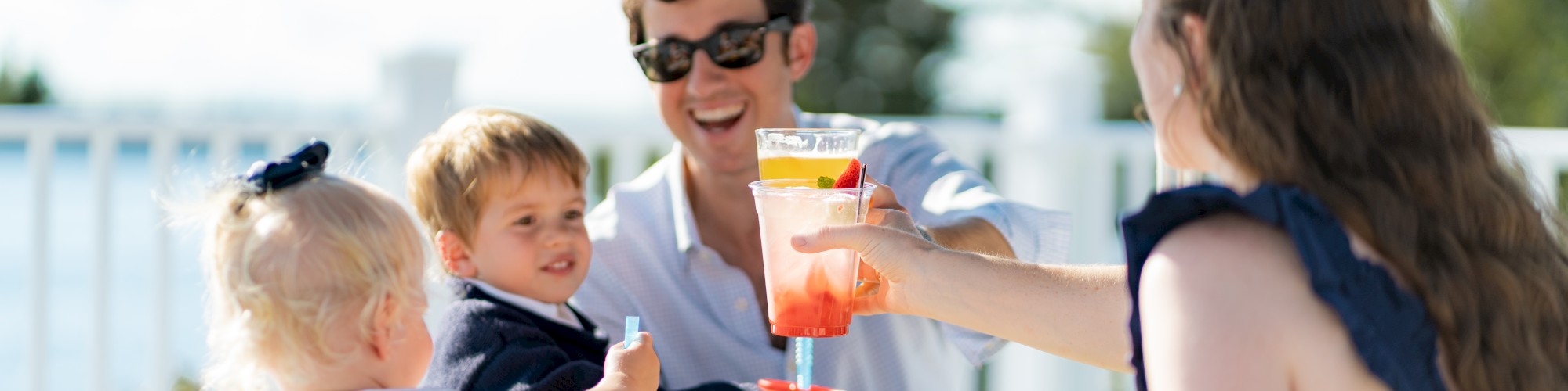 A family enjoys an outdoor meal together; two adults toast drinks while two children are at the table with food, creating a cheerful scene.