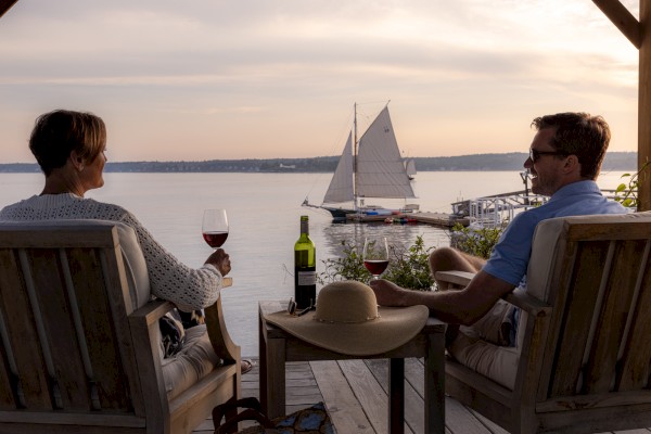 Two people relax with wine on a deck near water, watching a sailboat in the distance at sunset, creating a serene and scenic atmosphere.