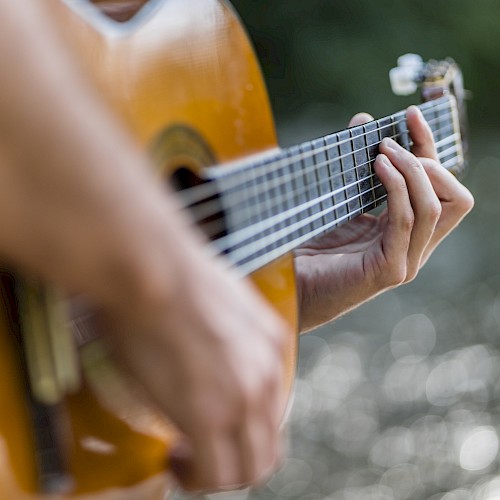 A person is playing an acoustic guitar with a blurred natural background.