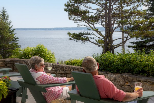 Two people relax in deck chairs, enjoying drinks and a scenic ocean view with trees in the foreground and a clear sky above.