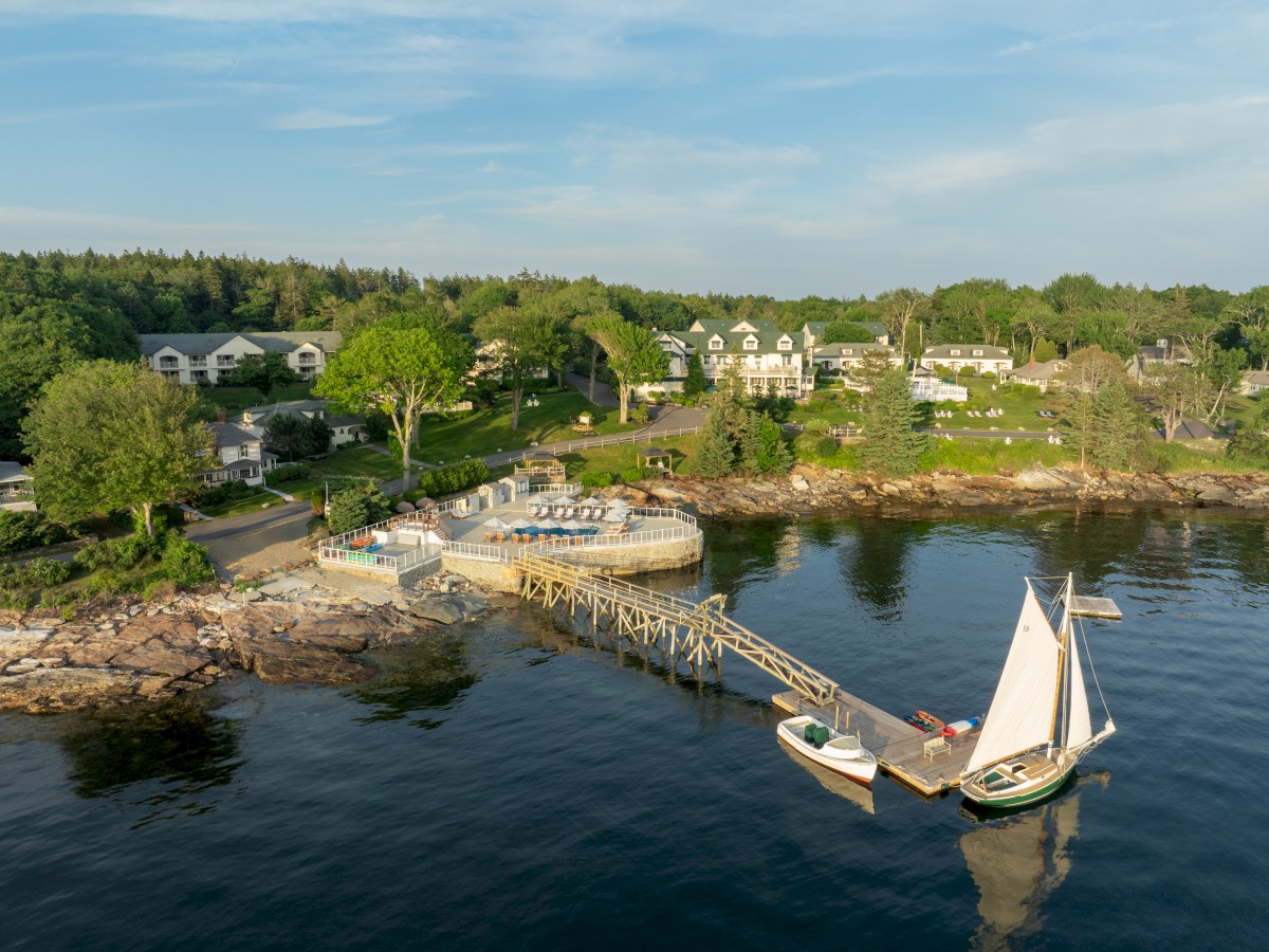 A coastal scene features a dock with boats, waterfront homes, trees, and a rock-lined shore under a clear blue sky ending the sentence.