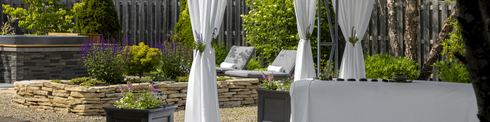 This image shows a serene garden with a gazebo, white curtains, lounge chairs, potted plants, and a bottle of water on a table, creating a relaxing space.