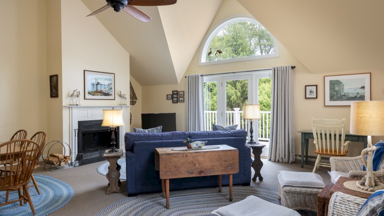 A cozy living room with a vaulted ceiling, blue sofa, wicker chairs, and a wooden coffee table. Large window with drapes and framed art.