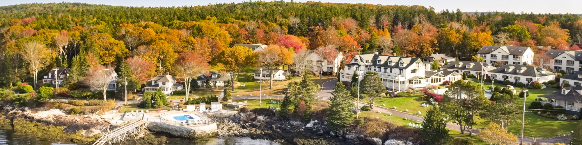 A coastal village with houses, autumn-colored trees, and a dock on calm water under a clear sky.