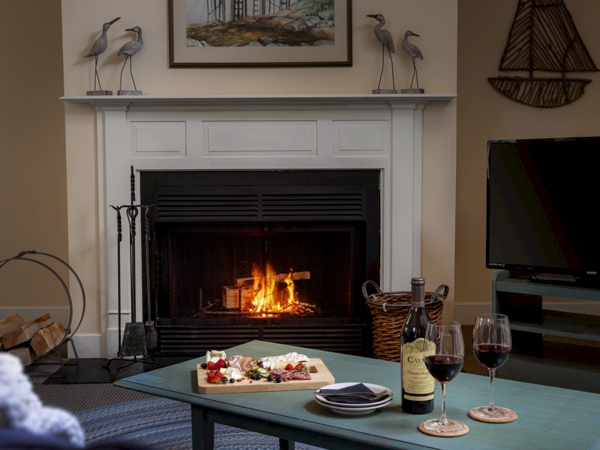 A cozy room with a lit fireplace, a table with wine and cheese, and a TV on the right. Decor includes bird statues and a sailboat art piece.