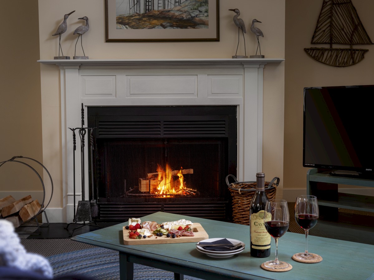 A cozy living room with a lit fireplace, cheese platter, wine, and TV. Decorative heron statues and a painting adorn the fireplace mantle.