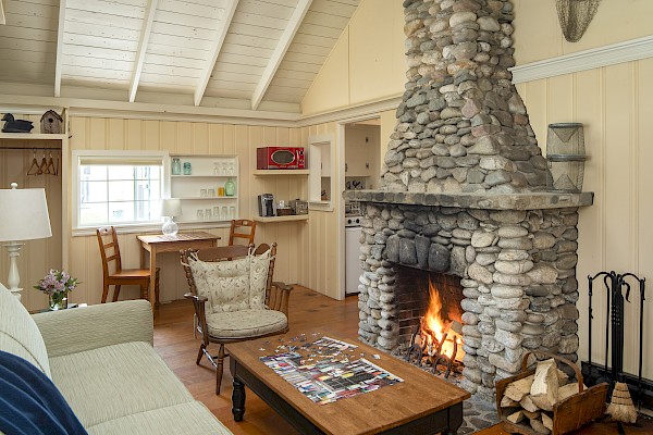 A cozy living room features a large stone fireplace, wooden ceiling, beige walls, and comfortable seating, with a dining area and small kitchen visible.