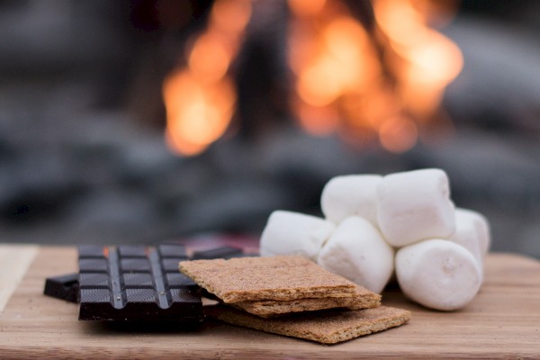 The image shows chocolate, graham crackers, and marshmallows on a wooden surface with a fire in the background.