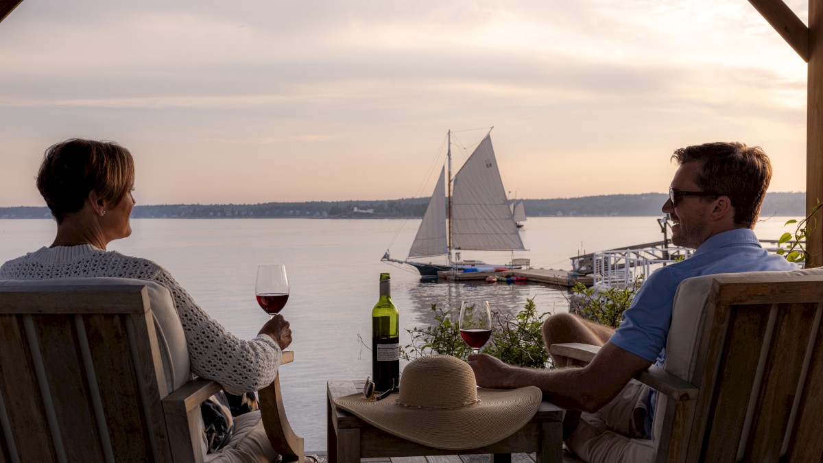 Two people sit on a deck with wine, watching a sailboat on the water at sunset. A hat and a wine bottle are on the table between them.