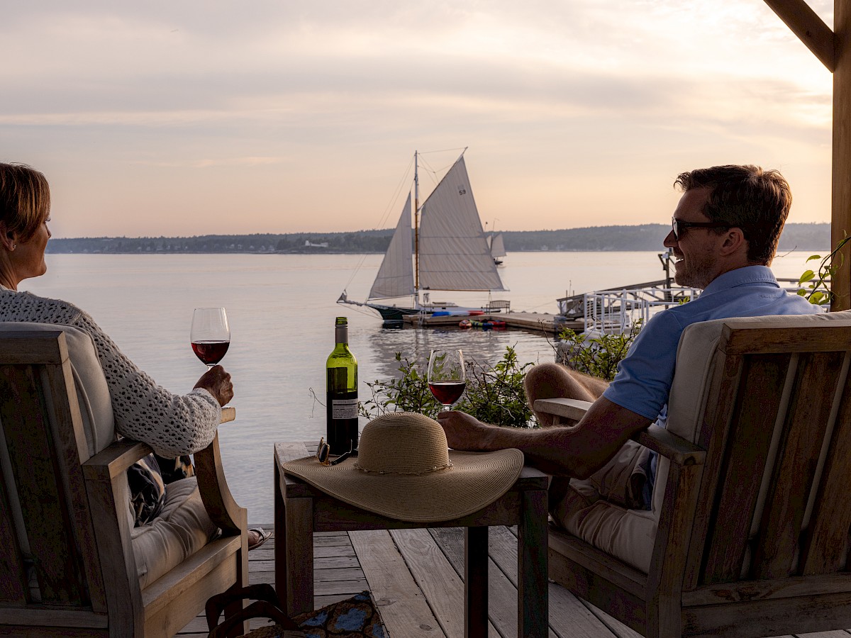 Two people sit on a deck with wine, watching a sailboat on the water at sunset. A hat and a wine bottle are on the table between them.