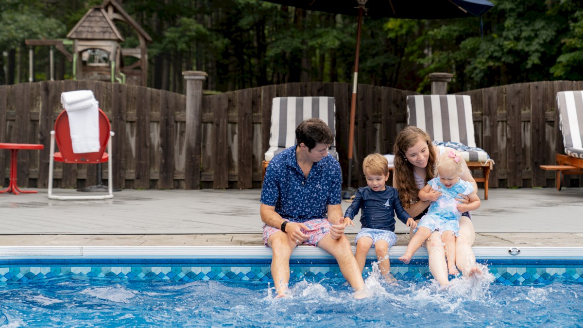 A family of four sits by a pool, dipping their feet in the water, with a fence and lounge chairs behind them.