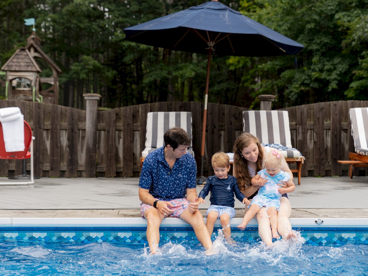 A family of four sits by a pool, dipping their feet in the water, with a fence and lounge chairs behind them.