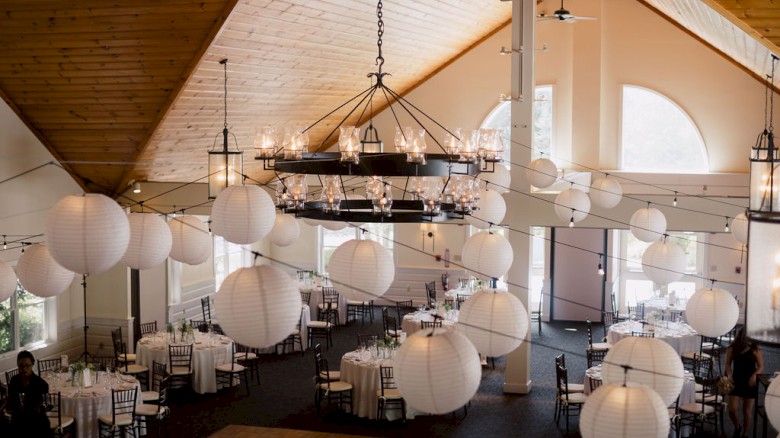 A decorated indoor event space with tables, chairs, chandeliers, and large hanging lanterns, creating a festive atmosphere.